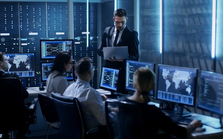 Na imagem aparece um departamento tecnológico com quatro funcionários em seus computadores e um executivo à frente.