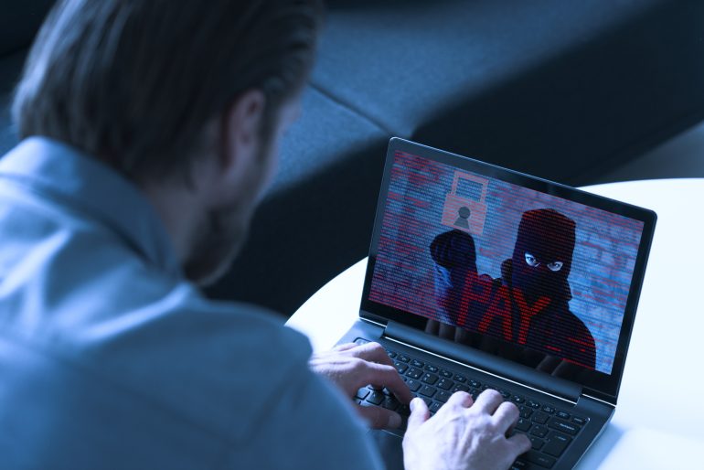 Homem digita em seu laptop. Uma imagem com um cadeado e o texto "pagar" aparece em sua tela. Talvez ele esteja criando um vírus de computador ou ransomware. Ou ele é a própria vítima.