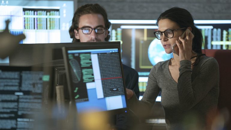 Homem e mulher que trabalha em um departamento cercado por monitores de computador, exibindo dados numéricos, gráficos em movimento e dados científicos.