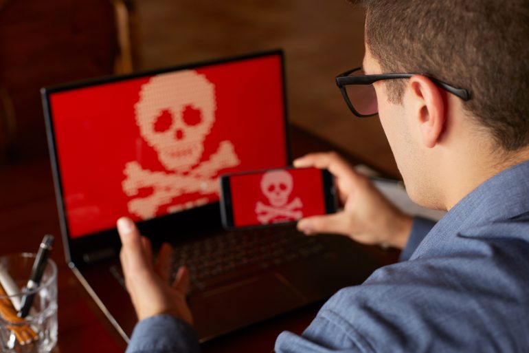 O homem senta-se perto do laptop com o telefone bloqueado e criptografado por spyware ransomware pedindo dinheiro. Laptop e smartphone infectados por vírus. Ossos cruzados de caveira vermelha assustadora na tela. Tema de segurança cibernética.