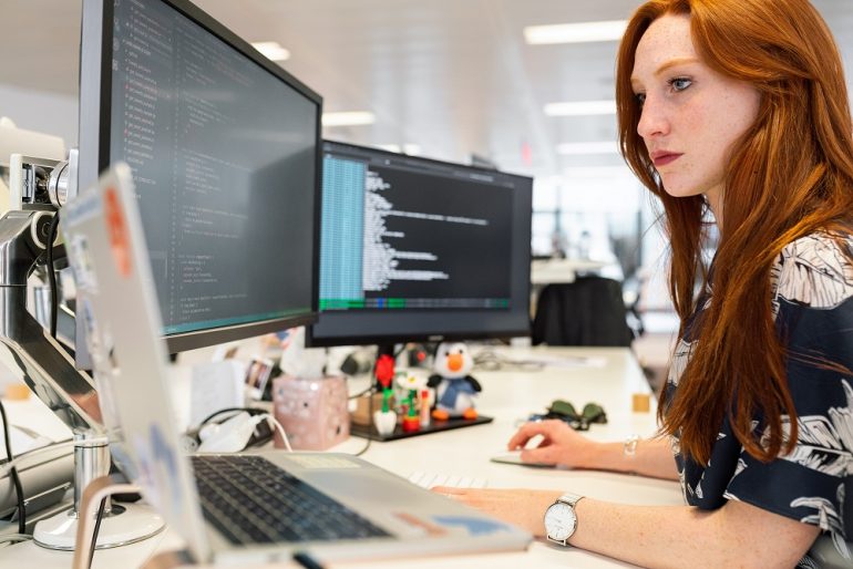 Imagem de mulher trabalhando no computador. Na frente dela possui duas telas com códigos descritos.