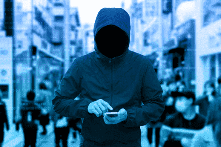 Imagem de um indivíduo com um blusão, encapuzado, andando pelas ruas com um celular em mãos.